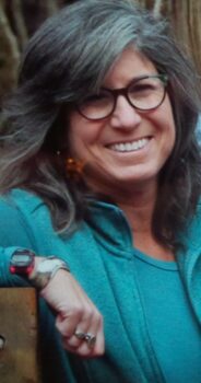 A woman with glasses, a smile, and long brown hair smiles. 