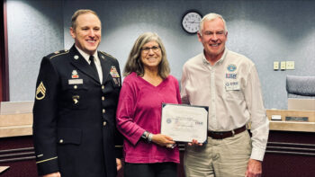 A smiling man in uniform, and another man from the USGR surround the recipient while she smiles and displays her award certificate.