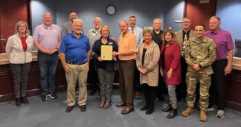 A group stands in the County Commissioners office and smiles. One holds a proclamation.