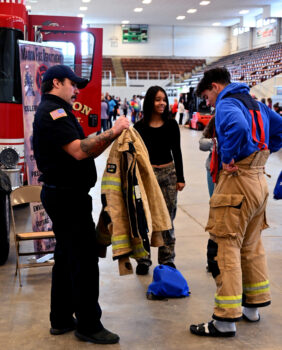 A firefighter helps a student try on the uniform while another student looks on. 