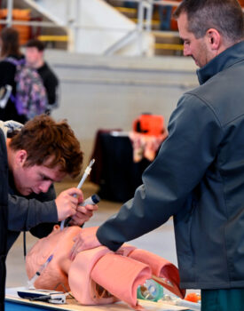 A student tries to insert tubes down a dummy's throat while an employee holds the dummy and provides instructions.