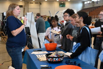 A woman talks behind a table to a group of teenage girls. 