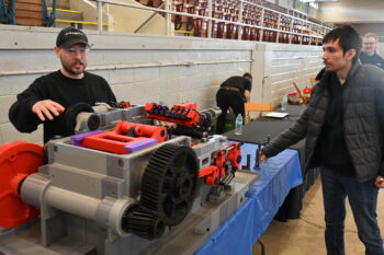 A student looks on while an adult gesture over a large plastic machine with many gears and moving parts. 