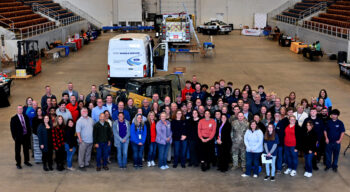 Dozens of exhibitors stand and smile in a group. Various pieces of equipment are around the coliseum, including a fire truck, highway patrol vehicle and more. 