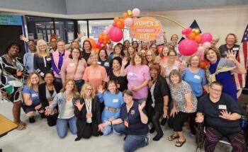 A large group of women smile underneath a balloon arch. 