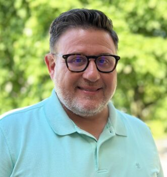 A white man with short brown hair, glasses, and a white beard. He is wearing a teal polo shirt and smiling.