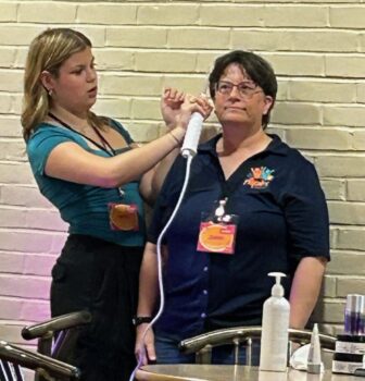 A young woman holds a white wand on the scalp of her mother. Skincare products are in the foreground.