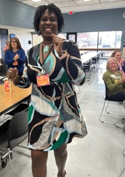 A black woman in a patterned dress smiles and dances to the front of the auditorium.