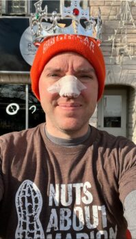 Daniel Bradshaw is a white man wearing a crown over his knit hat. He has tape over his nose to protect it while pushing the peanut across the ground. 