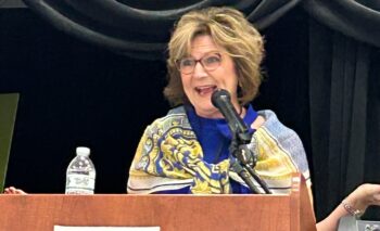 A woman with shirt brown hair, a blue dress and a blue and yellow scarf speaks behind a podium. 