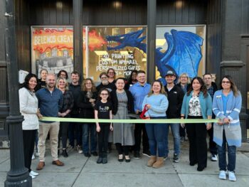 A group of 19 people smile. A ribbon is stretched in front and Maeghan Malone holds giant scissors poised to cut it. In the background, the store windows show a dragon with 'Belenos creations' in the fire it breathes. 