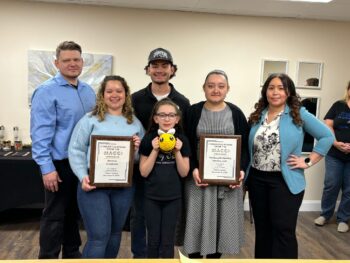 Two parents stand on either side of their four children: two adults, one teen and one child. Two of the daughters hold plaques from the Chamber. The youngest holds a toy bee.