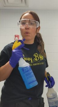 Ana is a Latino woman with long light brough hair in a pony tail. She is wearing glasses covered with safety goggles and gloves. She holds two spray bottles. 