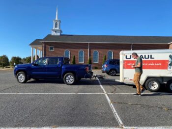 A truck pulling a small U-haul trailer is in the parking lot in front of a brick church with a white steeple. 