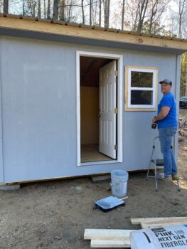 A man smiles while holding a drill. He is installing a window in a small shed with a door. Gray paint matching the exterior is in the foreground along with a bag of insulation and boards. 