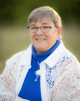 A white woman with short brown hair, black glasses, a smile, a royal blue turtleneck, and a white lace jacket. She also wears silver jewelry.