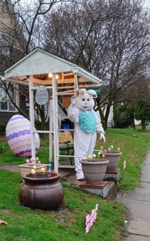 Tommy dresses up as the Easter Bunny and waves under the arch in front of his home. A large Egg and other decorations are behind him.