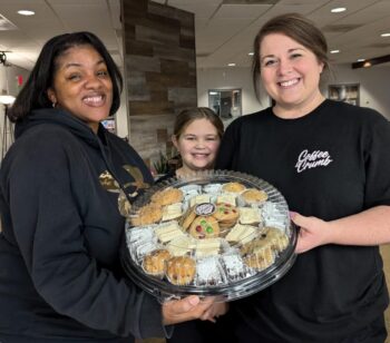 Two women smile while holding a tray of brownies, cookies, muffins and desserts. A young girl smiles between them.