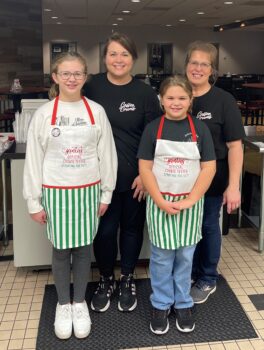 Two girls in aprons smile in front of their mother and grandmother.