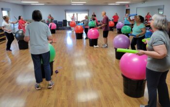 A group of seniors hold drumsticks and hit large, colorful exercise balls inside buckets. 