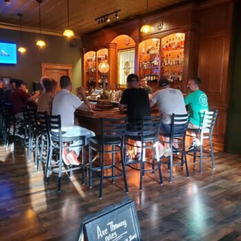 Photo of customers around the Kin & Kilt bar with wooden shelves of liquor behind the bartender.