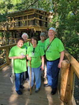 Three women and two men in matching Adventure Club shirts smile on a wooden bridge. Trees are in the background. 