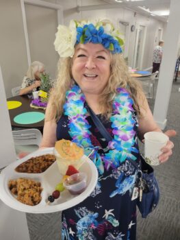 A woman with a lei a flowered headband smiles while holding food and a drink. 