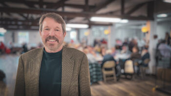 A man with light brown short hair and a trim beard and mustache smiles while wearing a dark green mock turtleneck and a brown checked suit jacket.