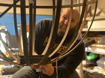 A smiling bald many with a brown beard smiles while holding a large metal orb. 