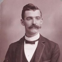 Frank Huber is a white man with short hair, a long curled mustache and a tuxedo.