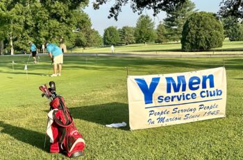 Golfers tee off while a banner advertises the Y-men Service Club and its motto: "People serving people since 1943."