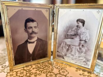 Two black and white portraits in gold frames. The man is a white man with a long, curled mustache and a tuxedo. The woman has dark hair in an updo and a white wedding dress. 