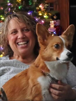 Nancy is a white woman with short brown hair and a wide smile holding her Corgi, a dog with reddish fur and a white streak.