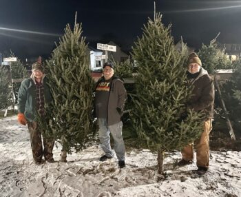 Three men smile while holding two Christmas trees. Many other trees for sale are visible in the background. 