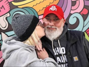 A woman with blonde hair, a knit cap and a sweatshirt kisses her husband on the cheek. He has a white beard, a red ballcap and an "Ohio Against the World" sweatshirt under a black vest.