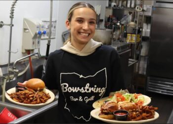 A young woman with blonde hair in a ponytail smiles while holding three platters of food in her arms.