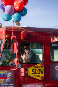 A man with sunglasses and a short beard waves while in a red truck with balloons tied to it. It says "Slow! Children crossing" and has a logo sharing that the food is free.