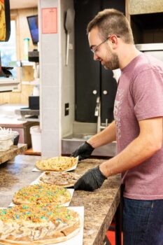 A young man with short hair and a trim bead and gloves makes dessert pizzas.