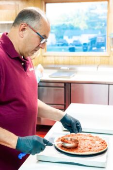 A man with glasses and gloves puts a tomato sauce on a pizza.