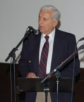 A man with gray hair and a suit speaks at a microphone in a color photo.