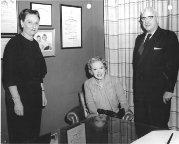 A black and white photo of a tall woman with a black suit and pearls and a tall man with white hair, a duit and glasses and a mustache on either side of a smiling, oung blonde woman with short, wavy hair and a light sweater and blouse. 