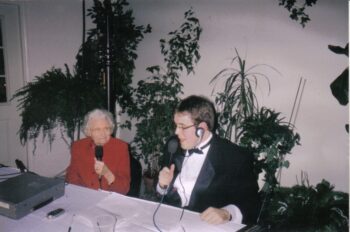 An elderly woman in a red suit and a young man in a tuxedo hold microphones. 