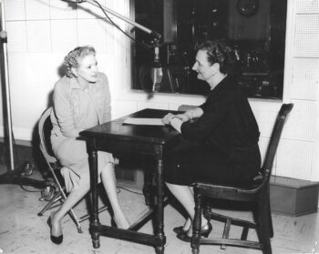 Two women sit at a small desk and chat with a large microphone between them in a black-and-white photo. 