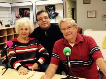 A woman, a younger man and an olderman smile in front of microphones.